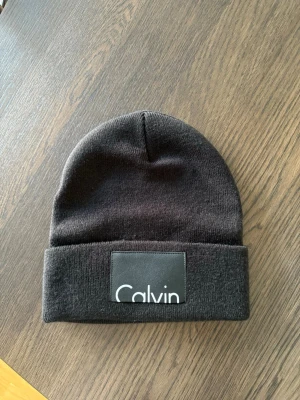 Svart mössa från Calvin Klein - Säljer en stilren svart mössa från Calvin Klein med uppvikt kant och en stor patch med logga framtill. Knappt använd. 