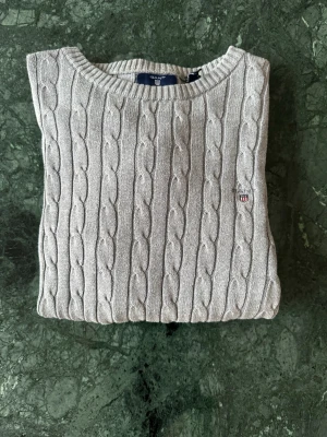 Grå kabelstickad tröja från Gant - Säljer en grå kabelstickad tröja från Gant med rund halsringning och diskret logga på bröstet. Tröjan har lång ärm och ribbade muddar vid ärmslut och nederkant. Perfekt för dig som gillar klassisk och stilren look.