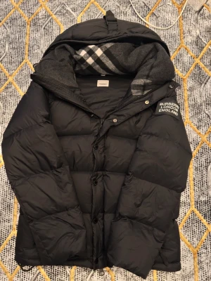 Svart pufferjacka från Burberry - Säljer en svart pufferjacka från Burberry med huva och klassisk logga på ärmen. Jackan har dragkedja och knappar framtill samt en mysig, vadderad känsla. Perfekt för kalla dagar och riktigt snygg till både jeans och sneakers.