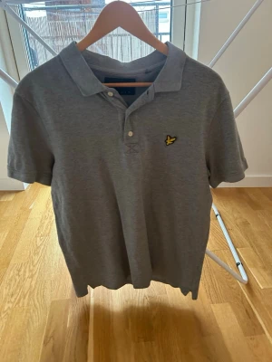 Grå pikétröja från Lyle & Scott - Säljer en klassisk grå pikétröja från Lyle & Scott med gul logga på bröstet. Tröjan har krage, korta ärmar och knappar framtill. Perfekt till jeans eller shorts för en clean och enkel stil.