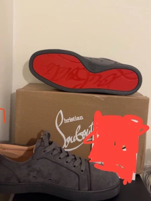 Grå Christian Louboutin sneakers mocka - Säljer ett par grå sneakers från Christian Louboutin i mocka med ikonisk röd sula och svarta nitar på tån. Skorna har snörning och rund tå, samt en platt sula. Perfekta för dig som vill sticka ut med exklusiv design och edgy detaljer.