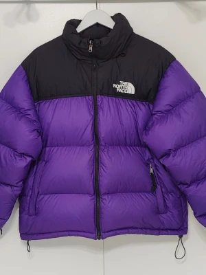 The North Face Nuptse 1996 Retro – Lila/Svart – Herr L - Säljer min TNF Nuptse 1996 Retro, herrmodell i storlek L, färg lila/svart. Köpt 2019 och äkta TNF, med 700-fill dun.  Jackan är använd men ren och fullt fungerande. Dunet är fortfarande puffigt och värmer som det ska.  Att känna till:  - Tvättlappen är svårläst p.g.a. tvätt (se bild). - Dragkedjan fungerar men är trög längst upp. Jackan går att stänga, men köparen bör vara medveten om detta.   Vänligen skriv vid frågor!