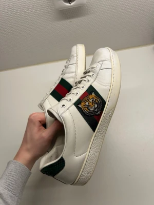 Gucci vita sneakers med tigerpatch - Gucci sneakers i vitt skinn med klassiska gröna och röda ränder på sidan och en broderad tigerpatch. Baksidan har metallicdetaljer i rött och grönt ormskinnsmönster med Gucci-logga. Skorna har snörning och platt sula, perfekt för dig som vill sticka ut.