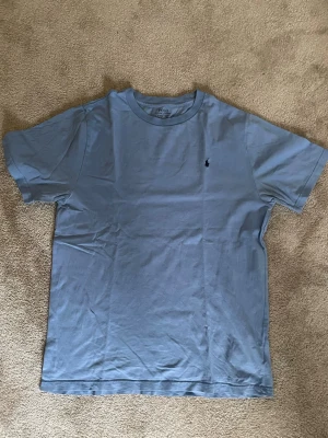 Ljusblå t-shirt från Polo Ralph Lauren - Säljer en ljusblå t-shirt från Polo Ralph Lauren i storlek XL (barn) som passar M/L i vuxen. Klassisk modell med korta ärmar och rund hals. Diskret logga broderad på bröstet. Tillverkad i mjuk bomull som känns skön mot huden. Perfekt till jeans eller shorts.