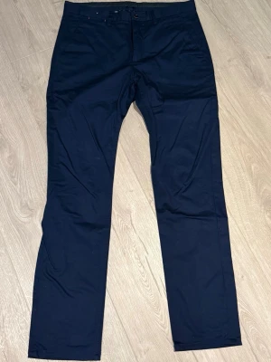 Mörkblå chinos med rak passform - Snygga mörkblå chinos med klassisk rak passform och diskreta fickor. Byxorna har bälteshällor och stängs med knapp och dragkedja. Perfekta för en clean och stilren look. Materialet känns mjukt och är troligtvis bomull eller en bomullsblandning.