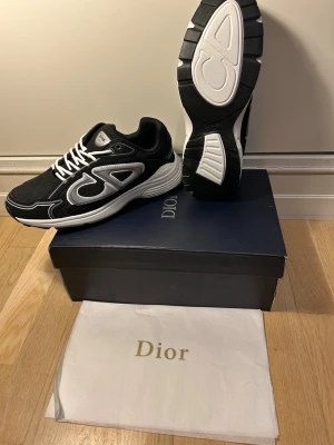 Svarta Dior sneakers med vita detaljer - Säljer ett par svarta Dior sneakers med vita och grå detaljer, tydlig Dior-logga på sidan och sula. Skorna har snörning och är tillverkade i textil och syntetmaterial. Snygg och modern design som sticker ut, levereras med originalkartong och dustbag.