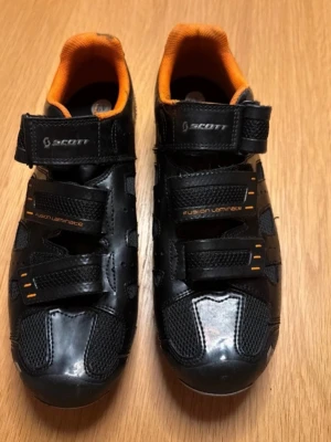 Scott Cykelskor MTB - Säljer ett par svarta cykelskor från Scott med orange insida och detaljer. Skorna har robust sula med grå och orange partier, kardborreband för enkel justering och meshpaneler för ventilation. Perfekta för dig som vill ha grymt grepp och snygg stil på cykeln. Kommer med shimano klossar