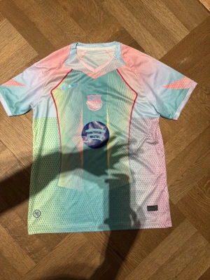 Nike FC Barcelona Lamine Yamal tröja - Säljer en FC Barcelona matchtröja från Nike med Lamine Yamal och nummer 10 på ryggen. Tröjan har pastellfärger i blått, rosa och mintgrönt, samt tryckta detaljer och Champions League-märke på ärmen. Tillverkad i Dri-FIT polyester för bästa komfort vid fotboll.