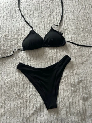 Svart bikini från H&M - Säljer en stilren svart bikini från H&M med trekantsbikinitopp och klassiska bikinitrosor. Topp med knyt i nacken och ryggen, båda delar i mjukt och stretchigt material. Materialet är polyamid och elastan, vilket gör den bekväm och följsam. Underdel storlek 34 S och överdel storlek 38 M