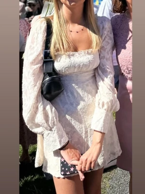 Vit kort klänning med puffärmar - Superfin vit kort klänning med puffiga långärmar och fyrkantig urringning. Klänningen har ett subtilt blommönster i tyget och markerad midja som ger en snygg siluett. Perfekt för sommarens alla tillfällen! Använd 1 gång.