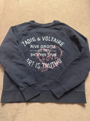 Svart sweatshirt från Zadig & Voltaire - Cool svart sweatshirt från Zadig & Voltaire i storlek XS med tryck i vitt och rosa på ryggen. Texten och hjärtat i taggtråd ger en edgy vibe. Tröjan har långa ärmar och är knappt använd!