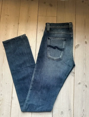 Blå Nudie Jeans Straight 27/34 - Säljer ett par klassiska blå jeans från Nudie Jeans med straight fit och snyggt slitna detaljer. Jeansen har fem fickor, ljusare tvätt på låren och bakfickor med Nudie-loggan. Passar dig som gillar tidlös stil och bekväma jeans.