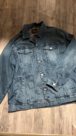 Blå jeansjacka från Dressmann 6XL - Snygg blå jeansjacka från Dressmann i slim fit-modell. Klassisk design med krage, bröstfickor med knappar och sidofickor. Jackan har långa ärmar och silverfärgade knappar framtill. Perfekt för dig som gillar en tidlös och cool look.