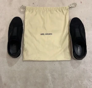 Axel Arigato skor - Snygga svarta sneakers från Axel Arigato i mocka och skinn med vit platt sula. Minimalistisk design med diskret logga i guld på sidan och elastisk detalj över vristen. Skorna levereras med original dustbag. Perfekta för dig som gillar stilrena och trendiga sneakers.