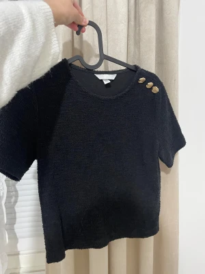 Svart kortärmad topp med guldknappar H&M - Svart kortärmad topp från H&M med rund halsringning och tre dekorativa guldknappar på ena axeln. Toppen har en mjuk, lite texturerad yta och rak passform. Perfekt för dig som gillar stilrena detaljer och enkel design.