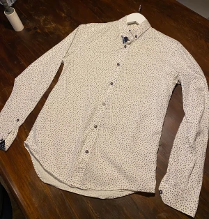 Blommig vit skjorta från Zara Man - Slim fit skjorta från Zara Man i vit med små blå blommor över hela tyget. Långärmad med mörkblå kontrasttyg och blommönster på insidan av manschetten. Klassisk krage och knappar framtill. Perfekt för både sommarfesten och skolan