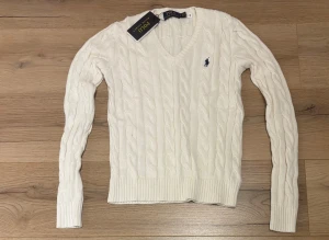 Vit kabelstickad tröja Polo Ralph Lauren - Vit kabelstickad tröja från Polo Ralph Lauren med klassisk v-ringning och marinblå broderad logga på bröstet. Tröjan har långärmad passform, ribbade muddar och är gjord i mjuk bomull. Perfekt för dig som gillar preppy stil och snygga detaljer.