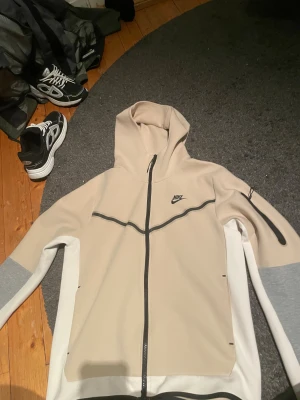 Beige Nike hoodie med dragkedja M - Snygg beige Nike hoodie i storlek M med svart logga på bröstet och hel dragkedja. Ärmarna har grå och vita detaljer, samt en praktisk ficka med dragkedja på ena armen. Materialet är mjukt och stretchigt, perfekt för chill eller träning.