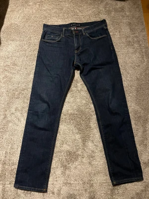 Mörkblå jeans från Tommy Hilfiger - Snygga mörkblå jeans från Tommy Hilfiger med klassisk femficksdesign och diskret logga på bakfickan. Jeansen har raka ben och normal passform, tillverkade i mjukt bomullsmaterial med lite stretch för extra komfort.
