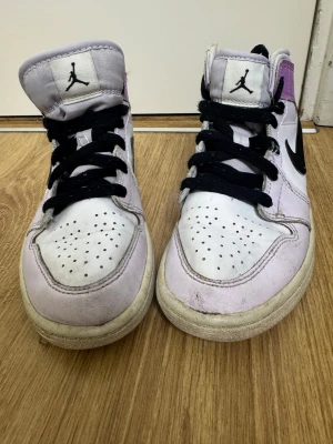 Nike Air Jordan 1 i vitt och lila - Säljer ett par Nike Air Jordan 1 sneakers i vitt och lila med svarta detaljer och klassisk svart swoosh. Skorna har högt skaft, svart snörning och perforerad tå. Materialet är skinn och textil, och de har den ikoniska Air Jordan-loggan på sidan.