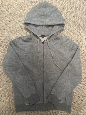 Grå hoodie med dragkedja och huva - En enkel grå hoodie med dragkedja framtill och snörning i huvan. Tröjan har långa ärmar, ribbade muddar och två fickor på framsidan. Perfekt för chill dagar eller när du vill vara extra bekväm. Materialet känns mjukt och är troligtvis bomull eller polyester.