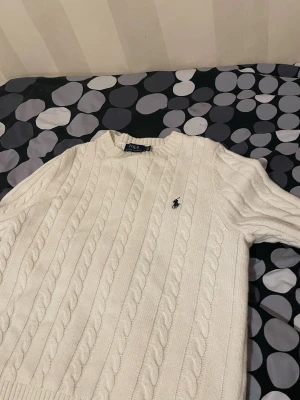 Vit kabelstickad tröja Polo Ralph Lauren - Vit kabelstickad tröja från Polo Ralph Lauren i storlek M. Klassisk design med rund hals och broderad marinblå logga på bröstet. Tröjan har långärmad passform och är gjord i mjuk bomull som känns skön mot huden. Perfekt för dig som gillar stilrena och tidlösa plagg.