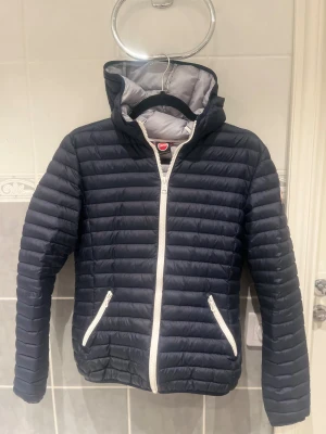 Mörkblå Colmar pufferjacka 14Y  - Snygg mörkblå pufferjacka från Colmar med vit dragkedja och två fickor med dragkedja. Jackan har huva och quiltad design som ger en cool look. Perfekt för dig som vill ha en lätt och varm jacka med stil. Colmar-logga på ärmen.