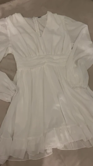 Klänning -vit med volang - Supersöt vit kortklänning med volangdetaljer längst ner. Materialet är lätt och V-ringad framtill och dragkedja bak. Köpte som extra klänning till studenten så den är oanvänd 