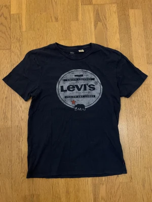 Mörkblå Levi's t-shirt med tryck - Snygg mörkblå t-shirt från Levi's i bomull med stort, grått tryck på bröstet och en liten röd stjärna. Klassisk rund hals och korta ärmar, perfekt för en chill och avslappnad stil.