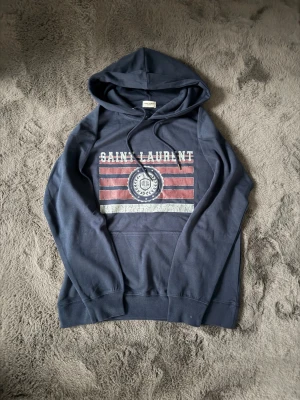Mörkblå Saint Laurent hoodie - Snygg mörkblå hoodie från Saint Laurent med huva och dragsko. Framsidan har ett tryck med röda och vita ränder samt ett SL-emblem i mitten. Tillverkad i mjuk bomull, perfekt för en chill och avslappnad stil.