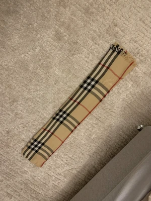 Rutig halsduk från Burberry London - Snygg halsduk från Burberry London med klassiskt rutigt mönster i beige, svart, vitt och rött. Halsduken har fransar i ändarna och är perfekt att slänga över axlarna för en cool och tidlös look. Materialet känns mjukt och lyxigt.