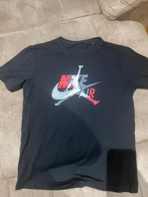 Svart Nike Air t-shirt med tryck - Svart t-shirt från Nike med stort Air Jordan-tryck i rött, vitt och grått på bröstet. Klassisk rund hals och korta ärmar. Perfekt för dig som gillar sportig streetstyle och vill ha en ikonisk look.
