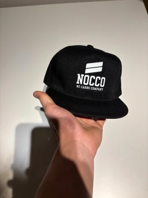 Svart snapback-keps från NOCCO - Snygg svart snapback-keps från NOCCO med vit broderad logga och texten 'NOCCO No Carbs Company' framtill. Klassisk platt skärm och justerbar baksida. Tillverkad i slitstarkt tygmaterial, perfekt för dig som gillar sportig streetstyle. Helt ny aldrig använd