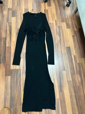 Svart lång stickad klänning med slits - Svart långklänning från SHEIN med ribbad stickad struktur, långärmad design och snygg v-ringning. Klänningen har en twistad detalj framtill och en slits på sidan för extra edge. Perfekt för dig som gillar minimalistisk stil med en cool touch.