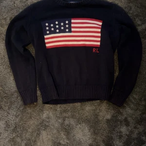 Mörkblå stickad tröja Polo Ralph Lauren - Mörkblå stickad tröja från Polo Ralph Lauren med amerikansk flagga i rött, vitt och blått på bröstet samt RL-broderi. Klassisk rund halsringning och ribbade muddar. Perfekt för dig som vill sticka ut med en ikonisk look.