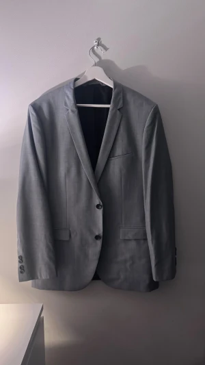 Blå kavaj från Hugo Boss - Snygg blå kavaj från Hugo Boss med klassisk passform. Kavajen har två knappar framtill, tre fickor och stilrena detaljer på insidan med röd accent. Perfekt för dig som vill ha en stilren och modern look.