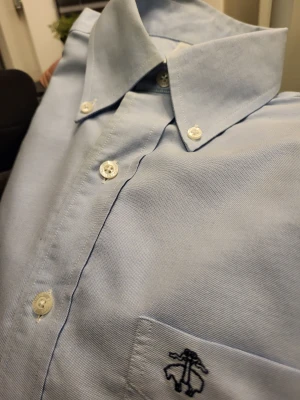 Ljusblå skjorta från Brooks Brothers - Klassisk ljusblå skjorta från Brooks Brothers i 100% supima bomull. Skjortan har button-down krage, vita knappar och en bröstficka med broderad logga. Långärmad och med stilren passform, perfekt för en clean och snygg look.