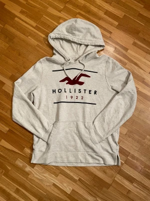 Grå Hollister hoodie med tryck - Snygg grå hoodie från Hollister med rött och svart tryck på bröstet. Klassisk huva med magficka framtill. Mjuk bomullsblandning som är skön att ha på sig.