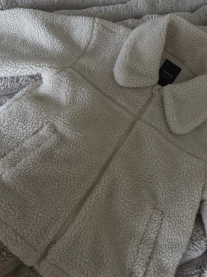 Beige teddyjacka från Name It - Mysig teddyjacka i beige från Name It med fluffigt material och stor krage. Jackan har dragkedja framtill och två sidofickor. Perfekt för dig som gillar chill och varm stil under höst och vinter.