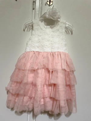 Vit och rosa volangklänning med blomdetaljer - Supersöt ärmlös klänning med vit överdel dekorerad med blomma  och fluffig rosa kjol i flera lager volangtyll. Klänningen har en stor rosett baktill och matchande blomaccessoar vid midjan. Det är för barn storlek mellan 2-3 år 
