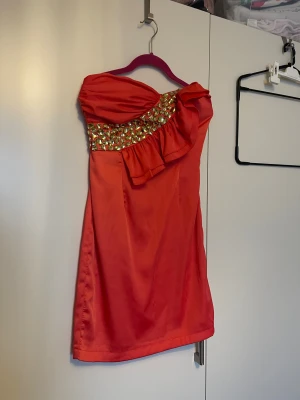 Röd axelbandslös kortklänning med volang - Snygg röd axelbandslös kortklänning med glittriga guldfärgade paljetter och volangdetalj över bysten. Klänningen har en slim passform och är tillverkad i ett glansigt syntetmaterial som ger en festlig vibe. Perfekt för dig som vill sticka ut med färg och detaljer.Använd bara några timmar 