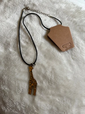Halsband med giraffberlock - Svart halsband med en cool giraffberlock i metall. Hängsmycket har en charmig design och halsbandet har justerbar kedja för perfekt passform. Perfekt accessoar för dig som gillar djur och unika smycken.