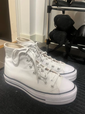 Vita Converse All Star platform - Säljer ett par vita Converse All Star med hög platåsula och klassisk rund tå. Skorna har snörning, silverfärgade öljetter och är tillverkade i textil med gummisula. Perfekta för dig som vill ha en clean och tidlös look med lite extra höjd. Är knappast använd.