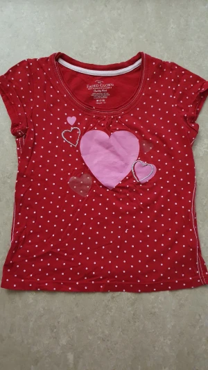 T-shirt - Söt röd t-shirt från Faded Glory med vita prickar och flera hjärtan i rosa och silver på framsidan. T-shirten har korta ärmar och är gjord i mjuk bomull, perfekt för en lekfull och färgglad stil.