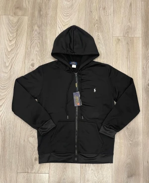 Svart hoodie från Polo Ralph Lauren - Svart hoodie från Polo Ralph Lauren med klassisk huva och snörning. Tröjan har dragkedja framtill, två fickor och den ikoniska vita Polo-loggan broderad på bröstet. Tillverkad i mjuk bomullsmix som är skön att bära. Perfekt för chill dagar och streetwear.