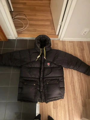 Svart dunjacka Fjällräven S - Säljer en svart dunjacka från Fjällräven med huva, gula snören och klassisk röd logga på bröstet. Jackan har stora fickor med knappar och dragkedja framtill. Perfekt för kalla dagar och riktigt snygg streetstil. Jackan har några små skador som ni kan se på bilden!går lätt att fixa till🫶🏻 kom gärna med egna priser också!