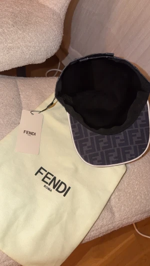 Svart Fendi keps med FF-mönster - Snygg svart keps från Fendi med ikoniskt FF-logomönster över hela kepsen. Kepsen har böjd skärm med vit kant och justerbar rem baktill. Tillverkad i textilmaterial och levereras med original dustbag och tagg.