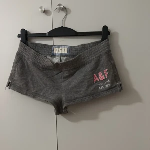 Grå shorts Abercrombie & Fitch - Snygga grå shorts från Abercrombie & Fitch med broderad rosa A&F-logga och vit text på ena benet. Modellen är lågmidjad med mjuk resår och tillverkad i bomull, perfekt för chill dagar. Klassisk och enkel design med kort passform.