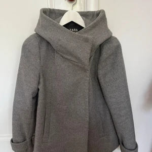 Grå kappa från Zara  - Snygg grå kappa med ullblandning från Zara Basic med krage och dold knäppning framtill. Jackan har långa ärmar, två fickor och är fodrad med svart tyg. Perfekt för kyligare dagar och ger en stilren look. Bra skick!! Dock knapparna är någorlunda lösa men det är enkelt att sy om om man skulle vilja