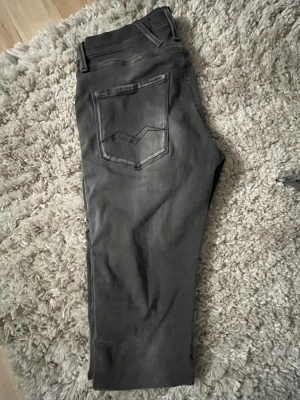 Replay Anbass  - Snygga Replay Anbass jeans i mörkgrå tvätt med slim fit. Klassisk femficksmodell med Replay-logga på fickan och coola detaljer på bakfickorna. Jeansen har dragkedja och knappstängning, och är gjorda i stretchigt jeanstyg för skön passform. Hör gärna av dig om du undrar något